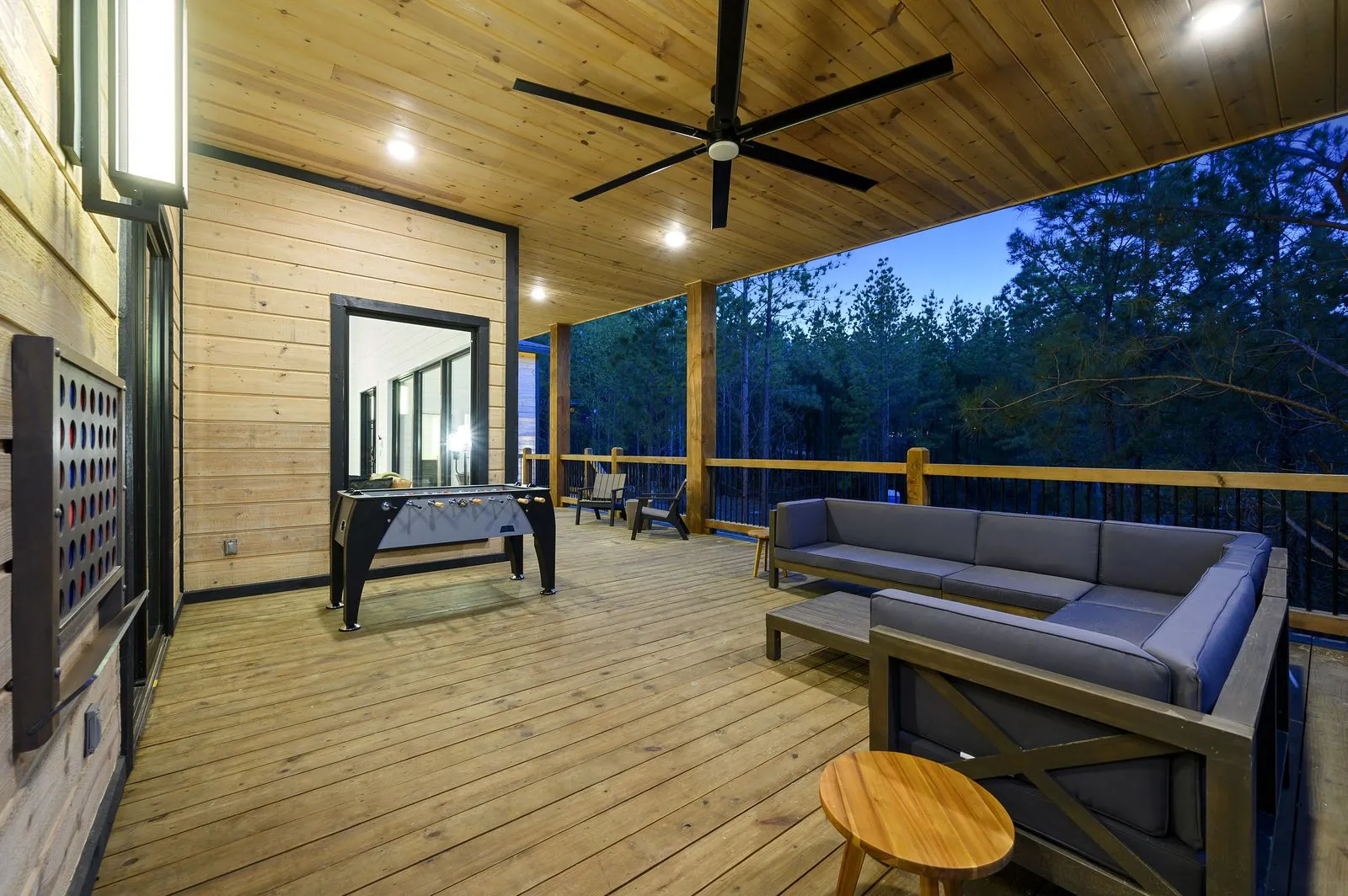 Covered deck at The Arcadia with outdoor sectional seating, ceiling fan, and forest views at dusk