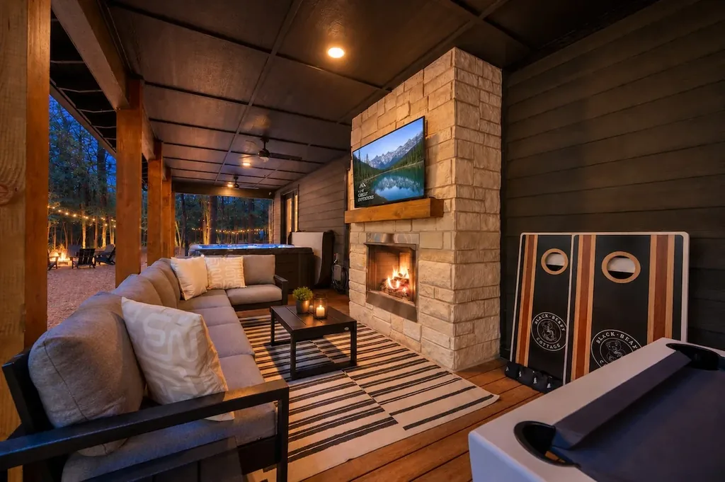 The Black-Beary — A covered outdoor living area with a sectional sofa, stone gas fireplace, mounted TV, and a hot tub and cornhole set nearby for evening use.
