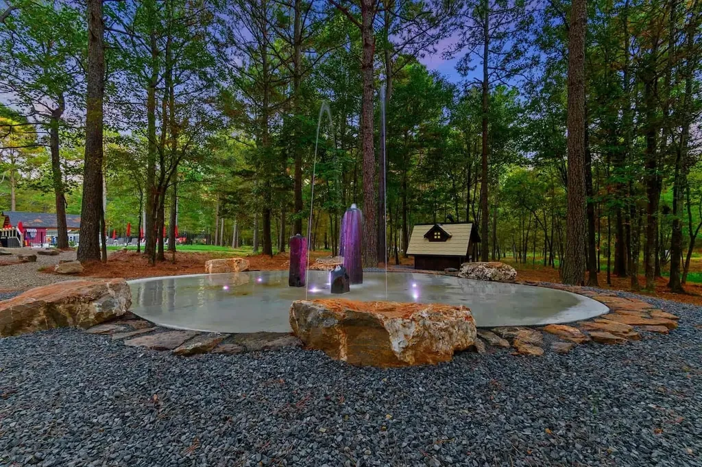 The Black-Beary — A shared splash pad with lit stone fountains sits in a wooded area of the resort, surrounded by boulders and gravel pathways. It is included at no extra cost for guests to use.