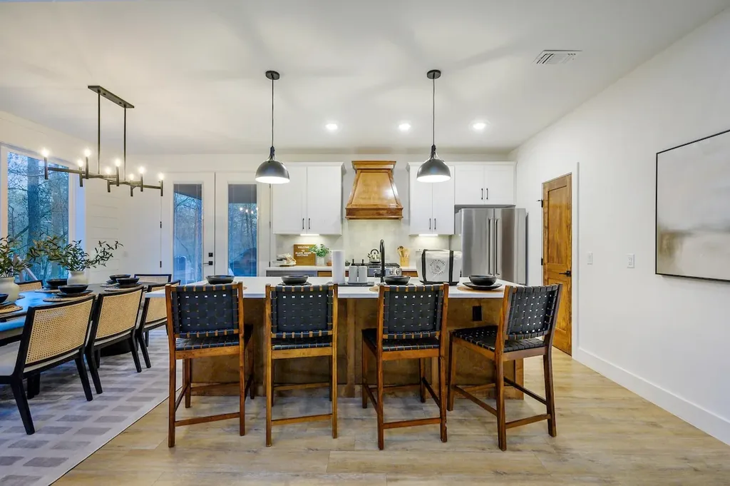 The Black-Beary — A large kitchen island with white countertop and four woven leather bar stools, backed by a white cabinet kitchen with a wood range hood and stainless refrigerator. Open dining area sits just beyond.