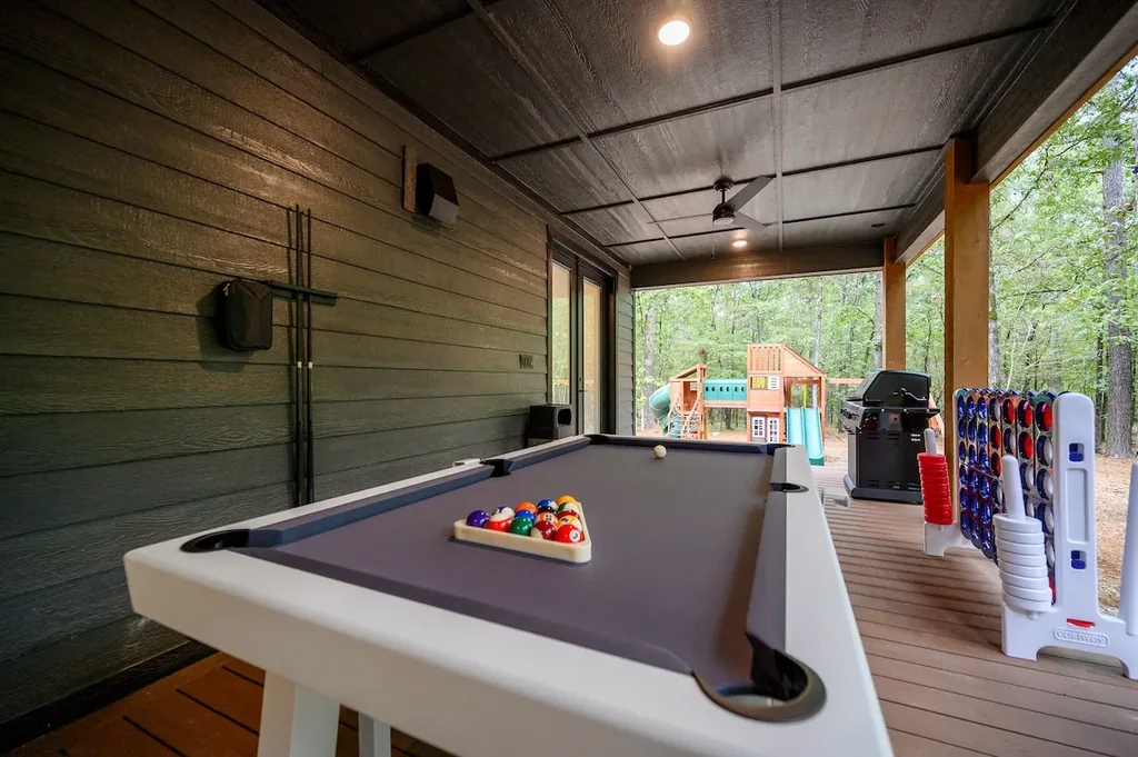 The Black-Beary — A covered patio with a private pool table, ceiling fan, and a gas grill, overlooking a wooded backyard with a children's play set.