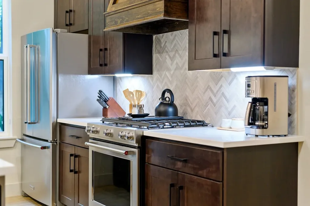 Dolce Far Nient — The kitchen features a stainless steel gas range and refrigerator, a chevron tile backsplash, and a drip coffee maker on the counter. Dark wood cabinetry with under-cabinet lighting lines the cooking area.