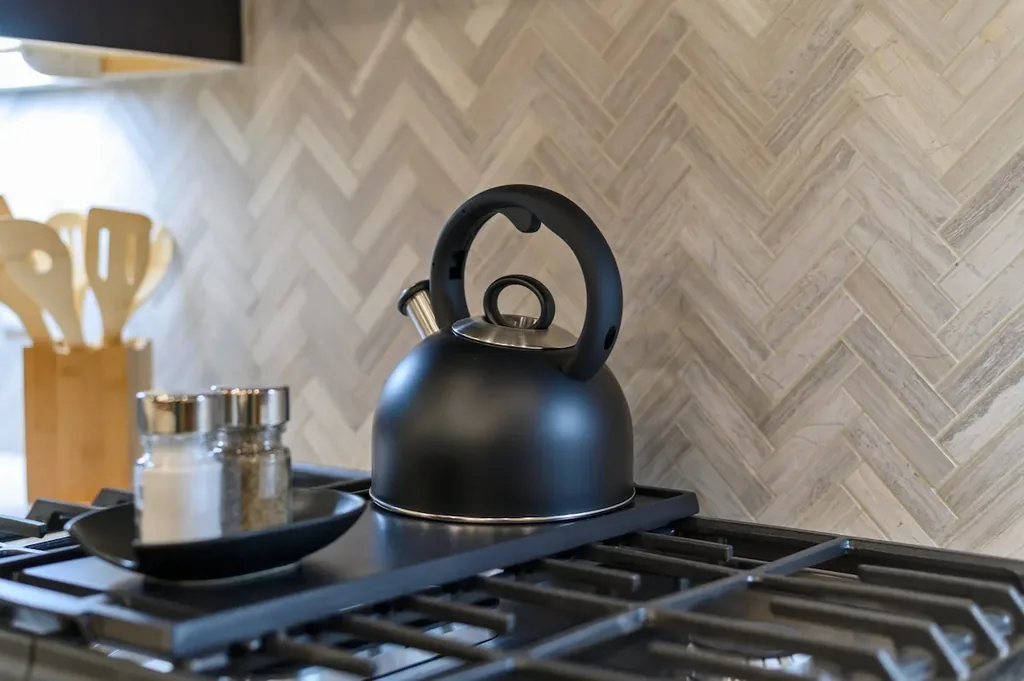 Dolce Far Nient — A stainless steel tea kettle sits on the gas stovetop, with salt and pepper shakers nearby and a herringbone tile backsplash behind.