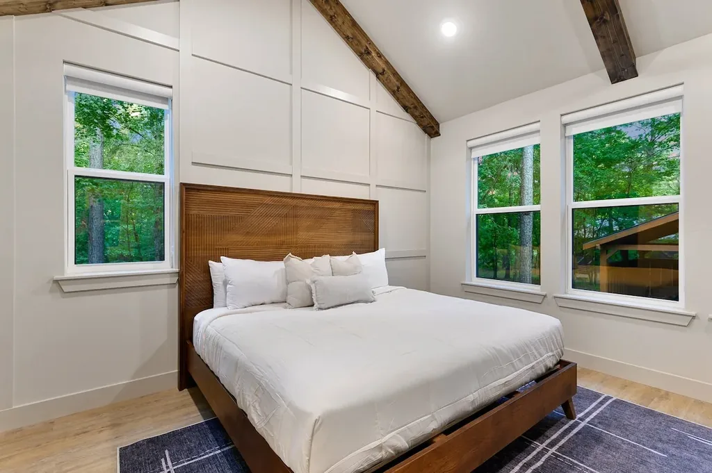 Dolce Far Nient — This bedroom features a wood-framed bed with a carved headboard beneath a vaulted, beamed ceiling. Large windows on two walls look out onto trees.