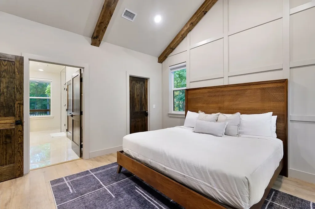 Dolce Far Nient — This bedroom has a king bed with a carved wood headboard and direct access to an en-suite bathroom. Exposed wood beams line the vaulted ceiling above, with a window offering a wooded view.