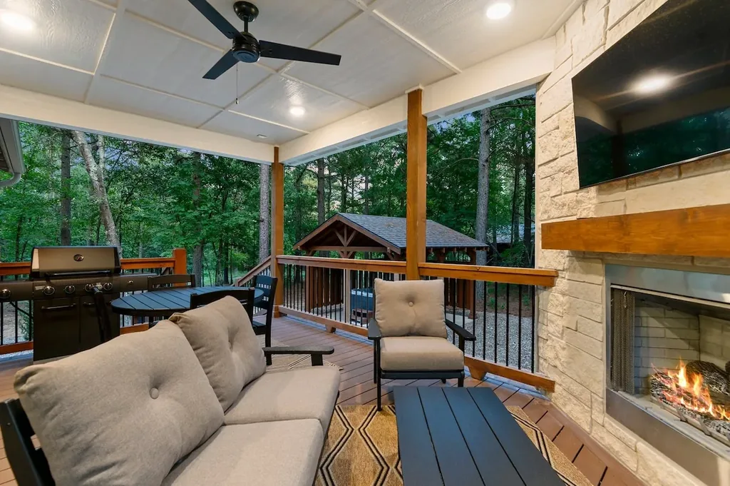 Dolce Far Nient — Relax by the stone fireplace on the covered patio, with a mounted TV, grill, and dining space steps away, all framed by tree views.