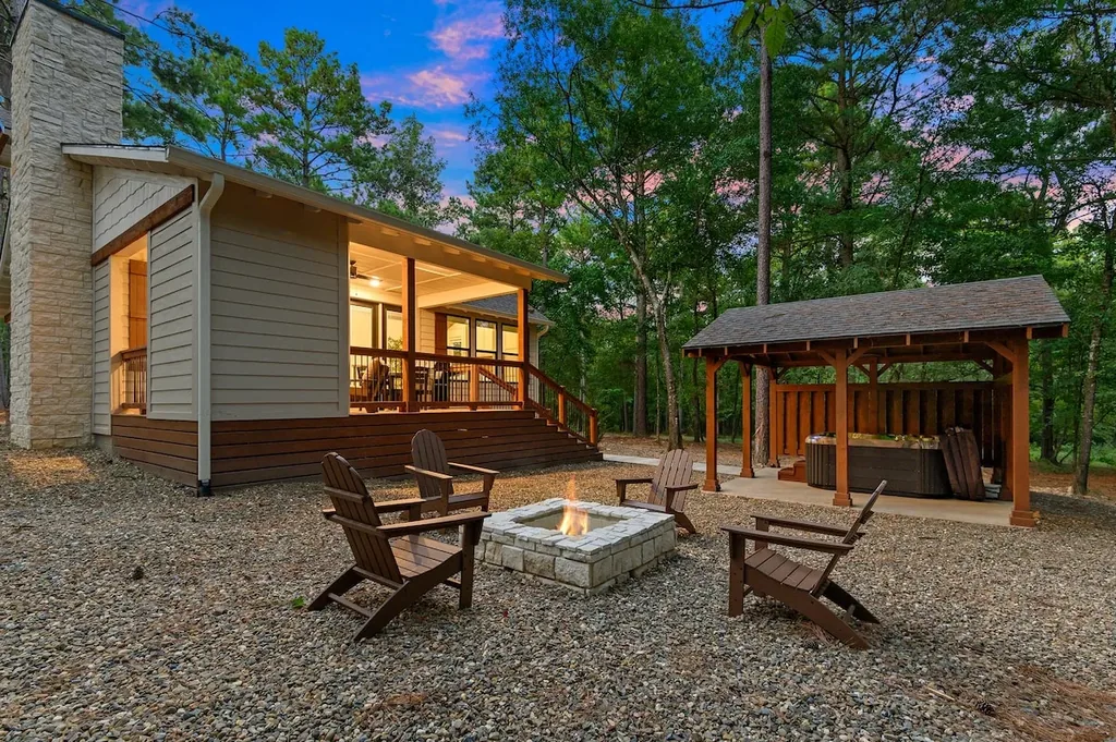Dolce Far Nient — A stone fire pit surrounded by four Adirondack chairs sits in the backyard, with a covered private hot tub under a wooden pavilion nearby.
