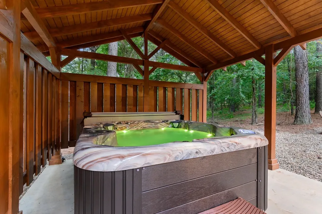 Dolce Far Nient — A private hot tub sits under a covered wood-beamed deck surrounded by trees, lit with color-changing lights.