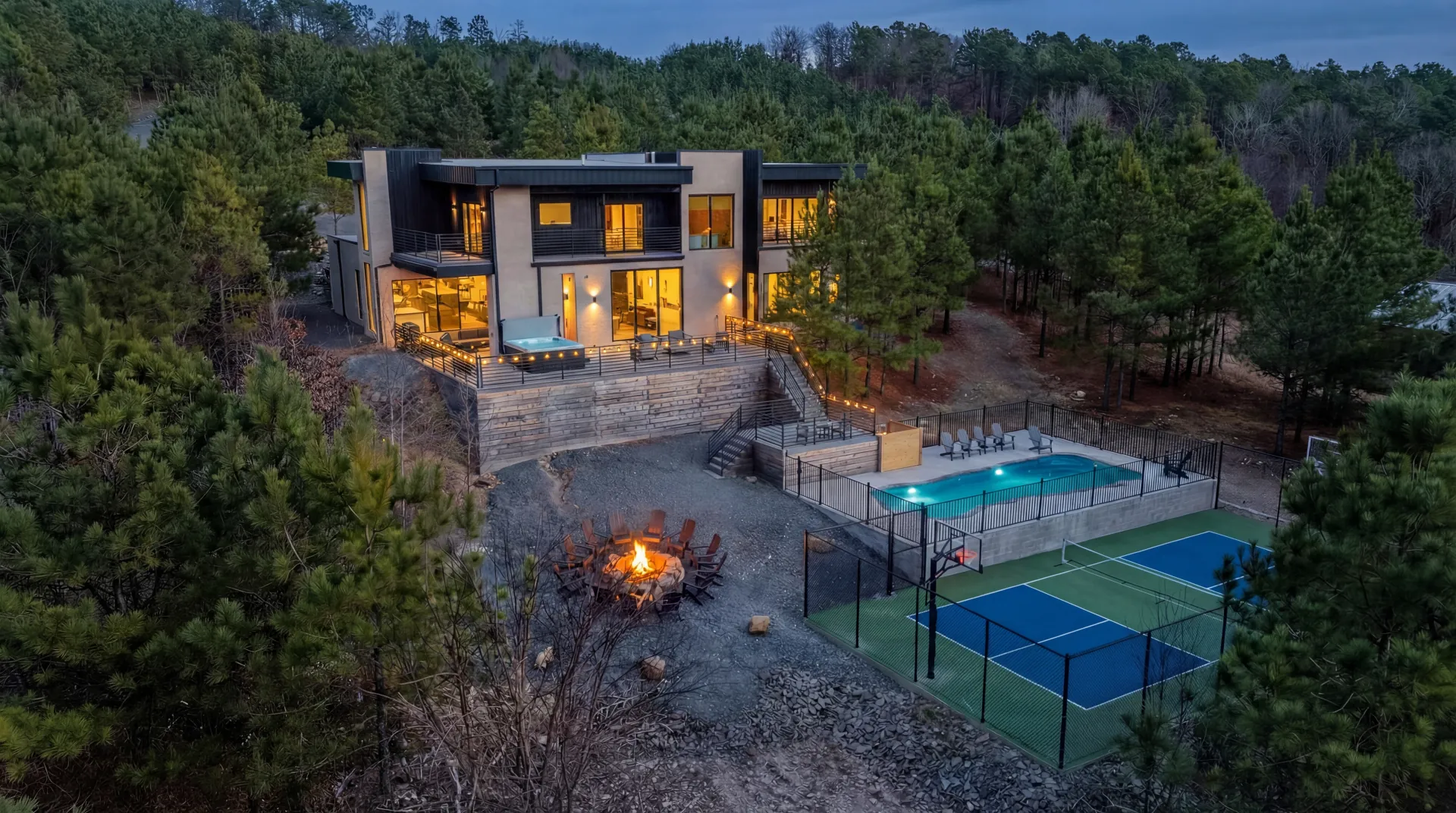 Twilight exterior of The Jade luxury cabin in Broken Bow with illuminated heated pool, hot tub, fire pit, and pickleball court surrounded by pine forest