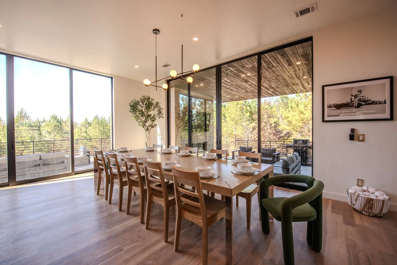 Formal dining room with long wooden table, designer green upholstered chairs, pendant chandelier, and floor-to-ceiling forest views