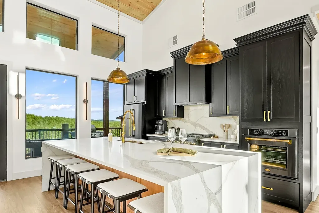 Chef's kitchen with marble waterfall island, black cabinetry, gold fixtures, and forest view