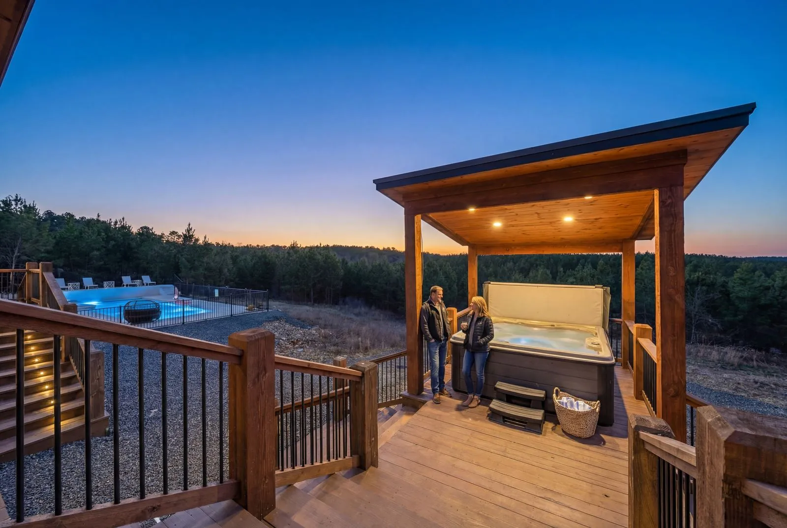 Couple beside the covered pergola hot tub at The Malibu at dusk, with the heated pool and forest sunset views below